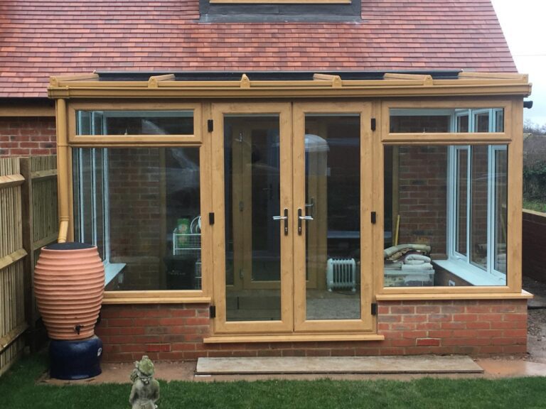 Stylish Irish Oak effect uPVC lean-to conservatory with large glass panels and a sloped roof, offering a bright and inviting extension that combines durability with a classic timber look.